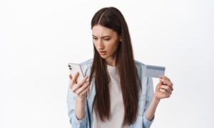 Online shopping. Brunette girl looks confused at smartphone screen, holding credit card, cant understand how register card to place an order, standing over white background.
