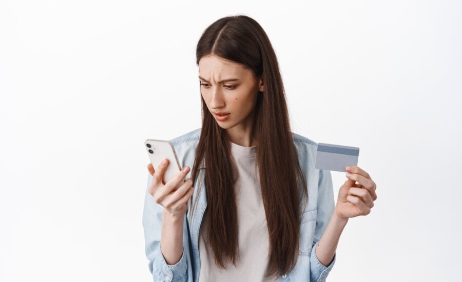 Online shopping. Brunette girl looks confused at smartphone screen, holding credit card, cant understand how register card to place an order, standing over white background.
