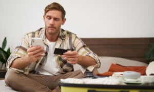 Angry man holds credit card and mobile phone, sits next to clothes in suitcase, has problems with paying online.