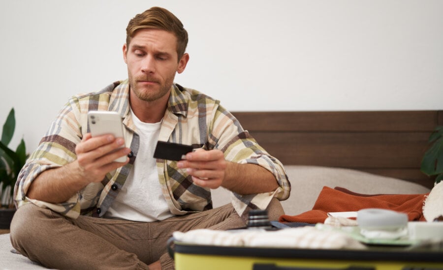 Angry man holds credit card and mobile phone, sits next to clothes in suitcase, has problems with paying online.