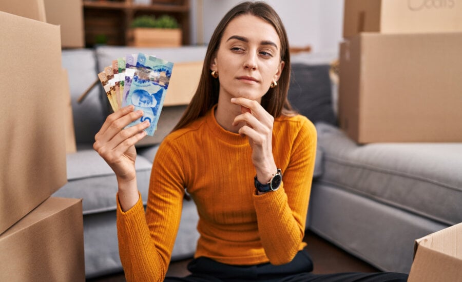 Woman sitting on the floor at new home holding canadian money serious face thinking about question with hand on chin, thoughtful about confusing idea