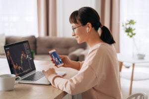 Smiling business woman trader analyst looking at laptop monitor, holding smartphone, wearing earphones. Investor broker analyzing indexes, trading online investment data on stock market graph at home,