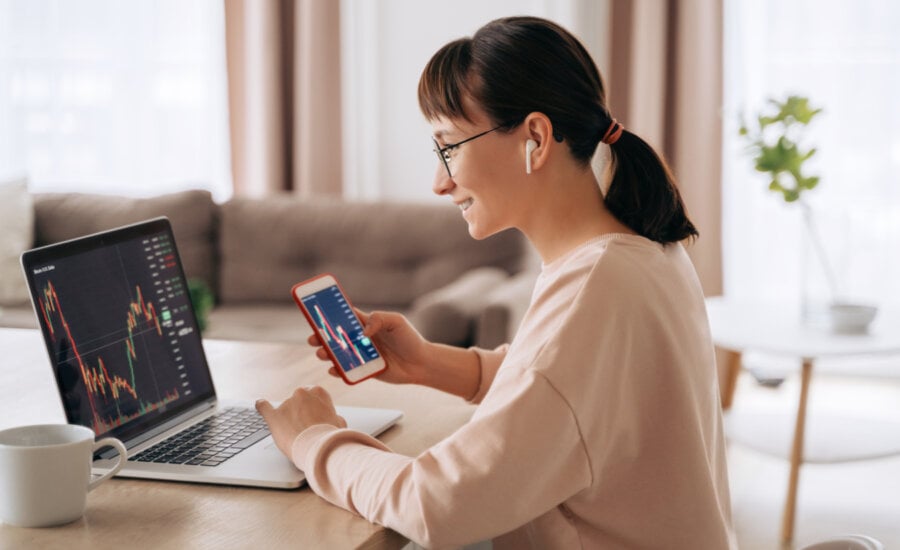 Smiling business woman trader analyst looking at laptop monitor, holding smartphone, wearing earphones. Investor broker analyzing indexes, trading online investment data on stock market graph at home,