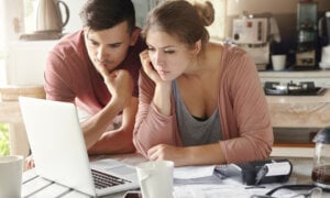 Serious man and woman sitting at kitchen table in front of open laptop computer, looking at screen with concentrated expression, focused on paying utility bills online. Family budget and finances
