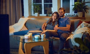 Loving young couple, man and woman sitting on sofa in evening at home,, hugging, cuddling and watching tv together. Concept of family, leisure time, relaxation, childhood and parenthood