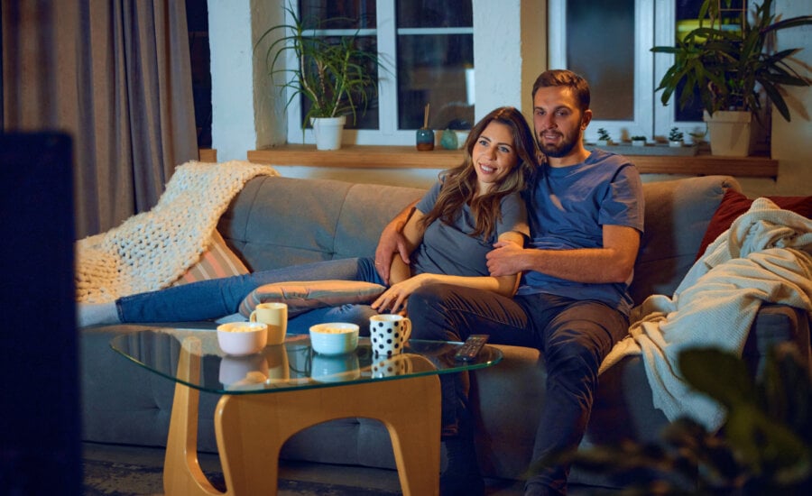 Loving young couple, man and woman sitting on sofa in evening at home,, hugging, cuddling and watching tv together. Concept of family, leisure time, relaxation, childhood and parenthood